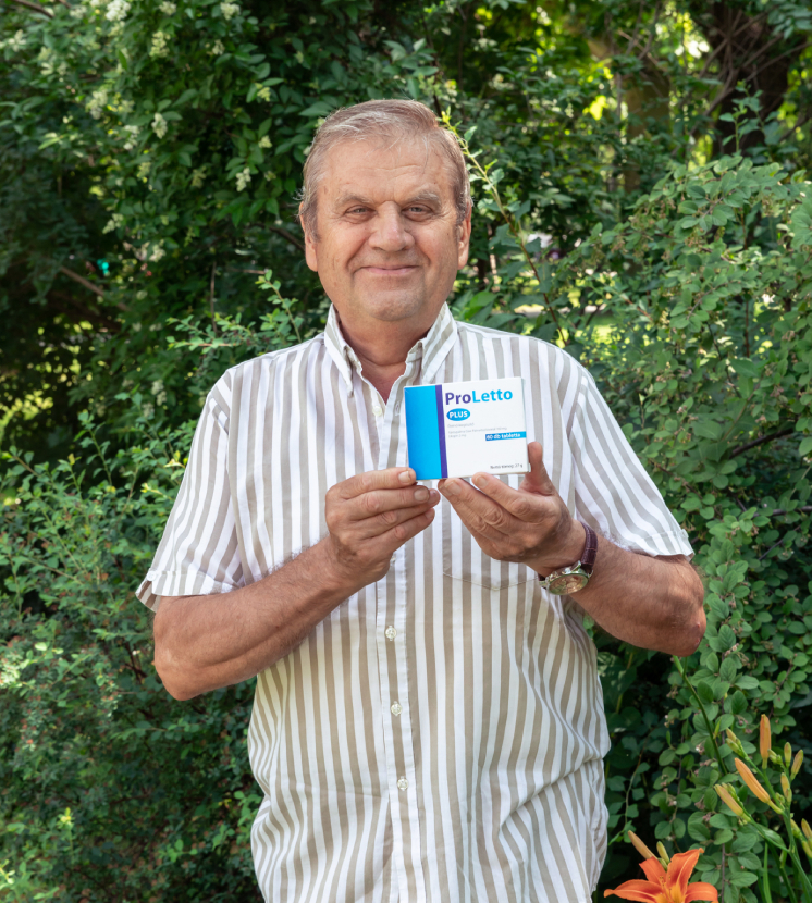 Natural Pharmaceuticals ProLetto® Plus Mosolygó idős férfi kék pulóverben kutyájával a természetes háttér előtt ülve.